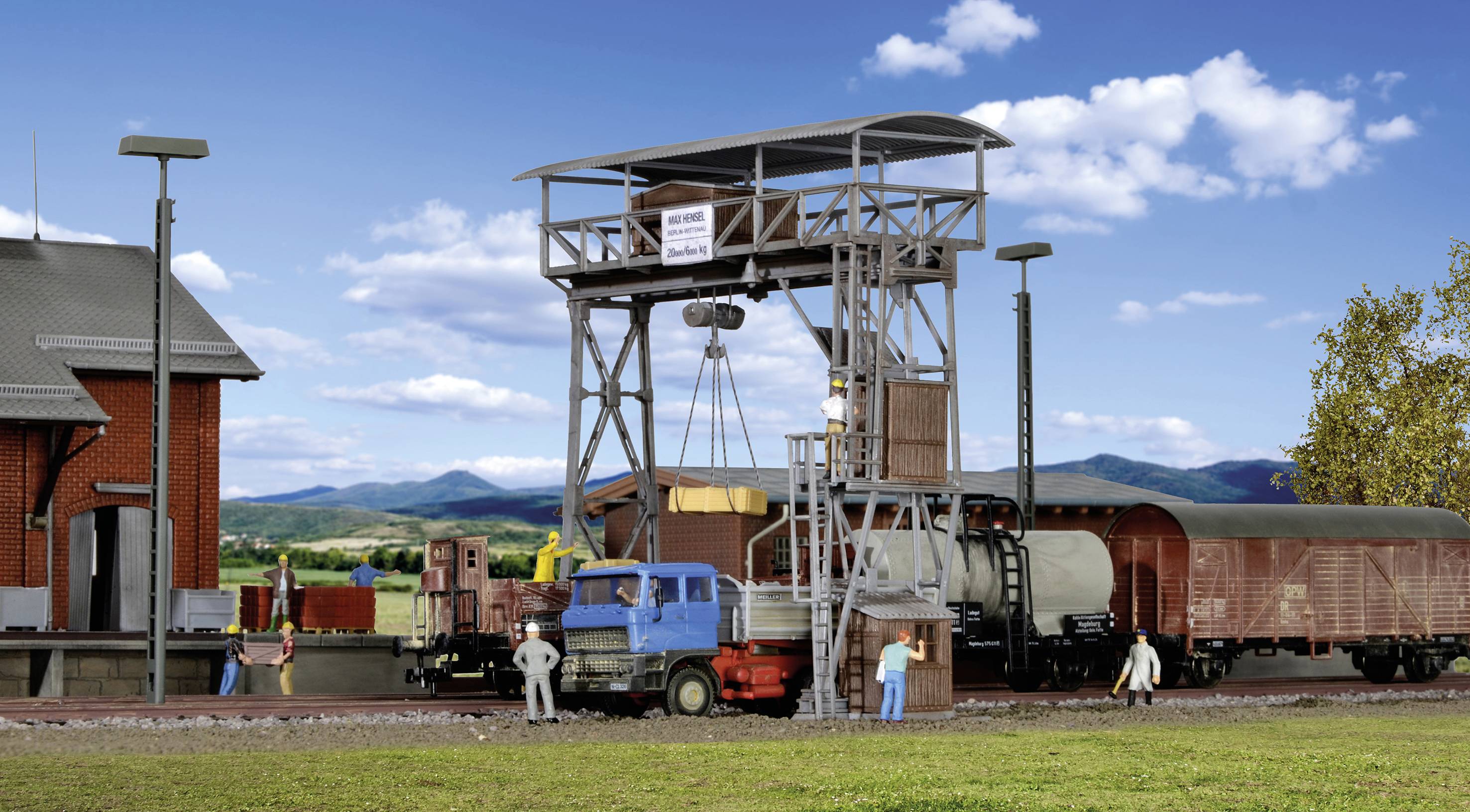 A railway loading ramp with multiple wagons and workers operating a crane. The background depicts a rural landscape with rolling hills.