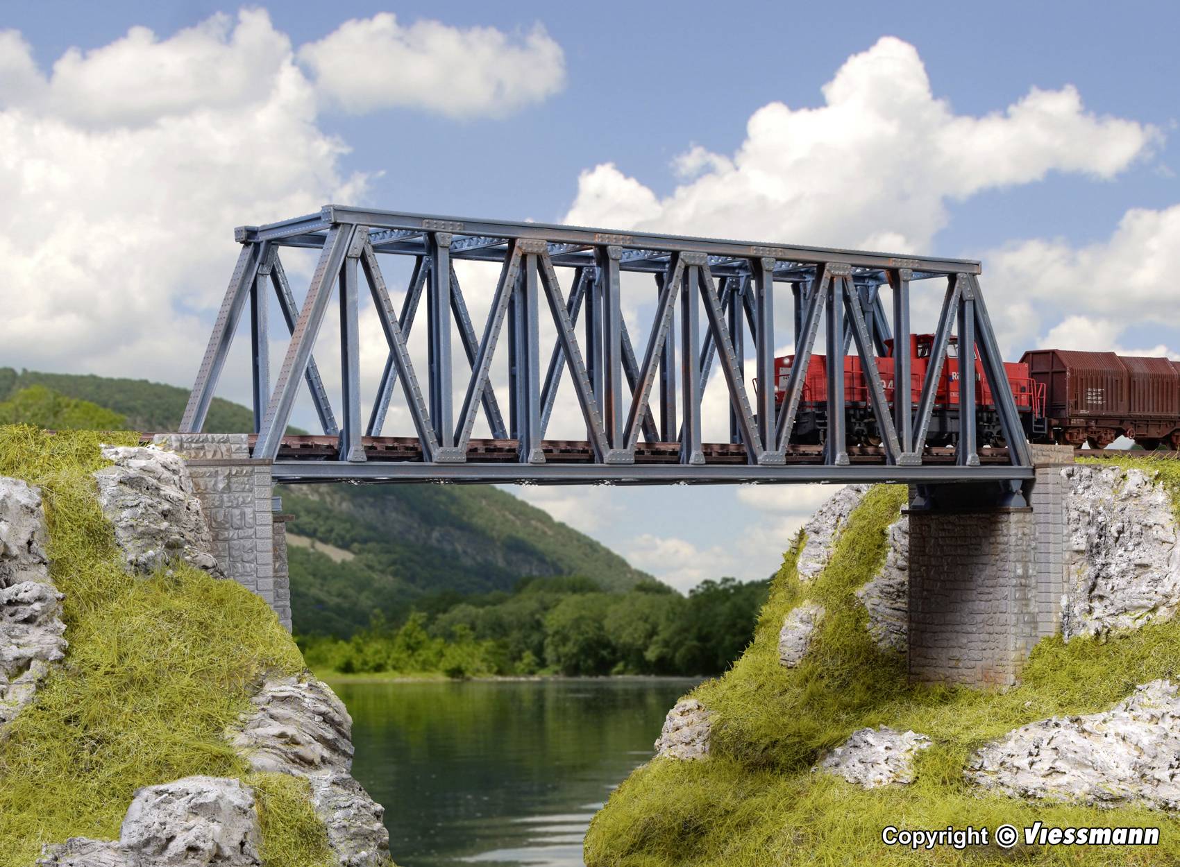 'A red train travels across a metal bridge spanning a river, surrounded by green hills and a blue sky.'