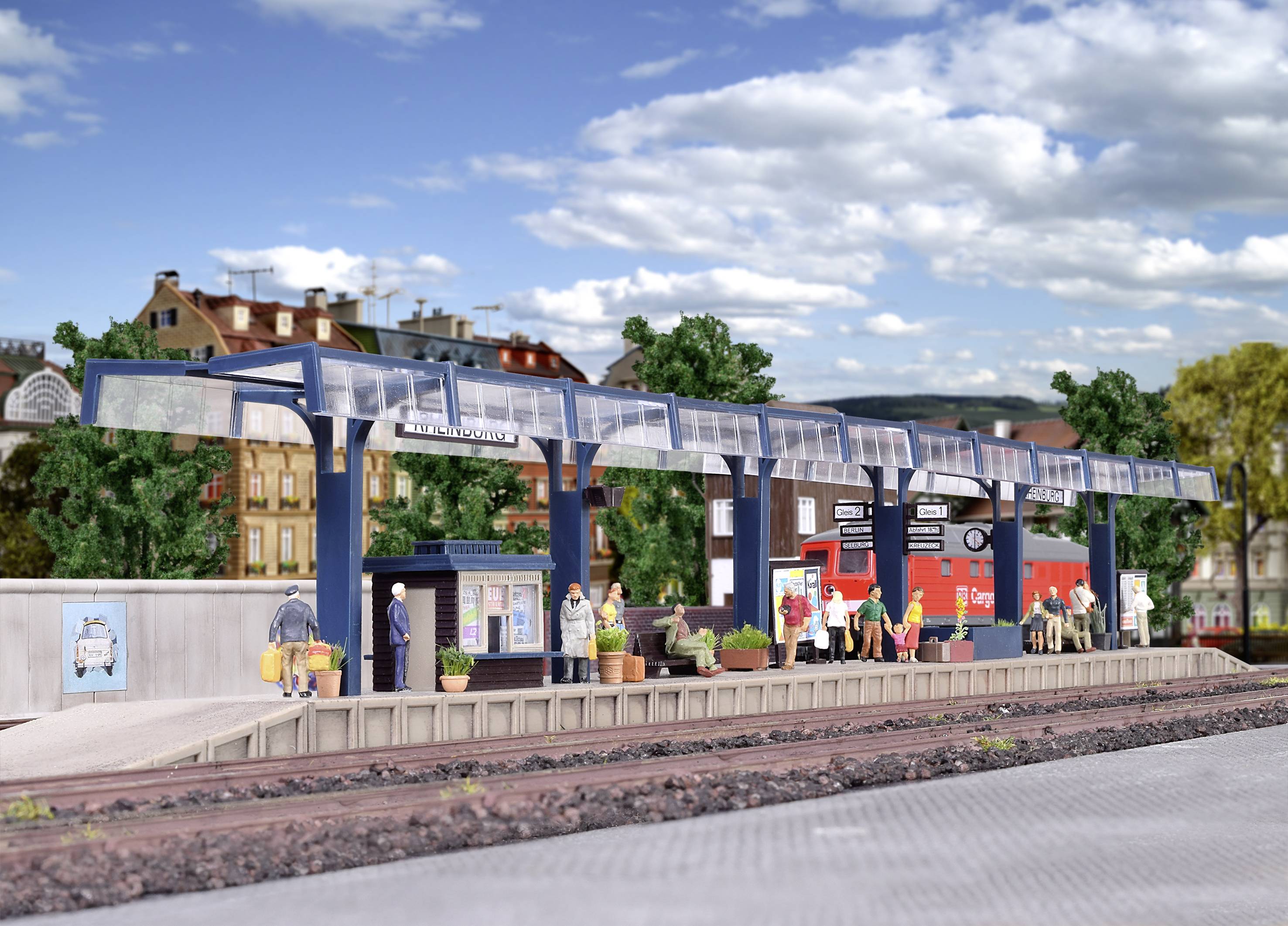 A model railway station with waiting figures on the platform. In the background, buildings and a blue sky can be seen.