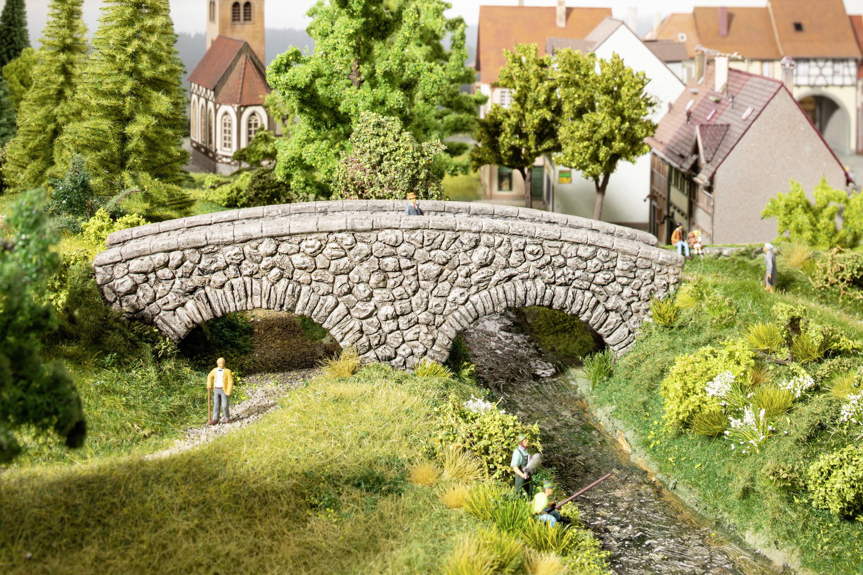 A diorama depicts a stone bridge over a small river, surrounded by trees and houses, with people walking and fishing.