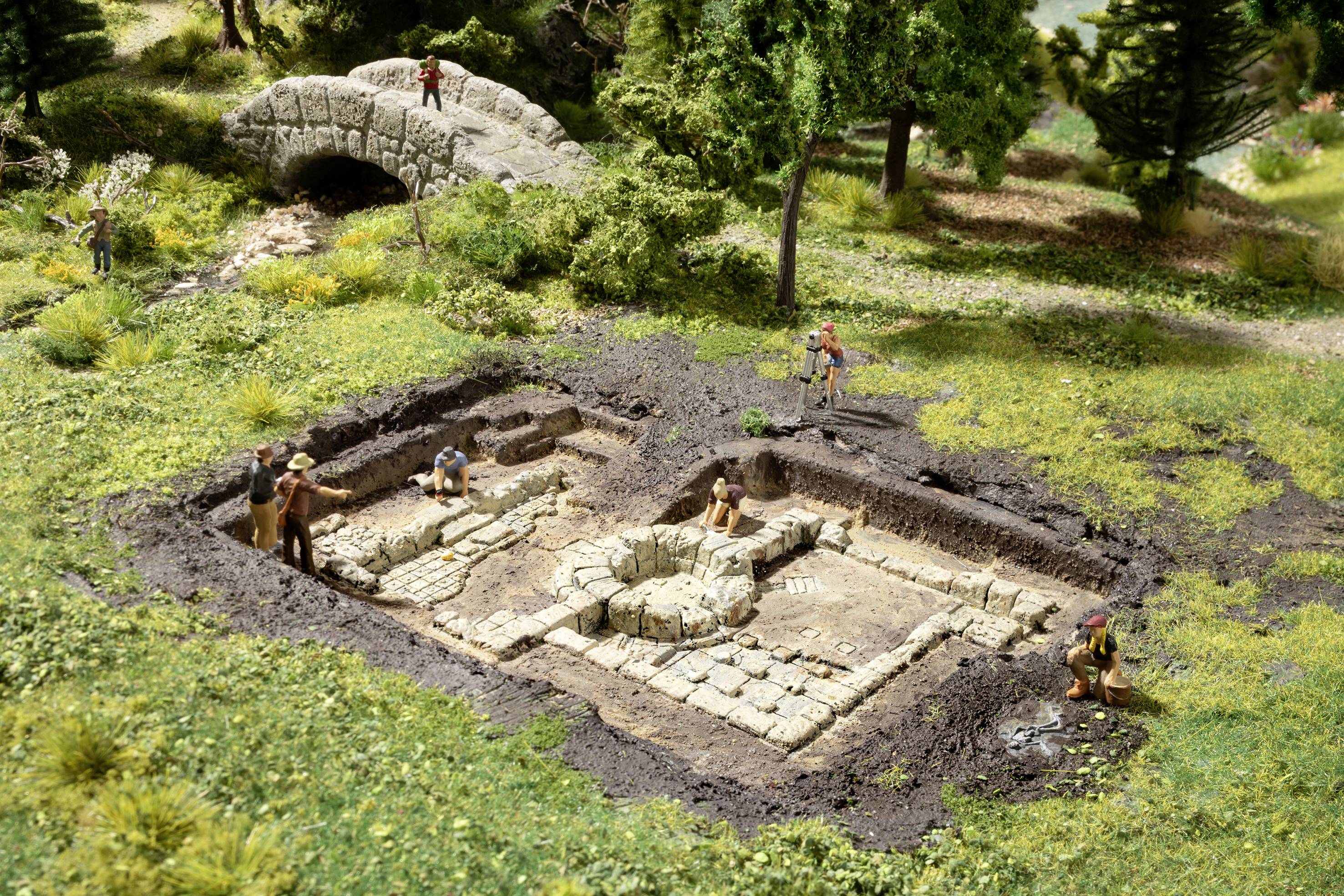 A miniature scene depicts an archaeological excavation site with figures working in an excavated area; a stone bridge is visible in the background.