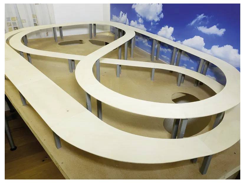 'Model race track on a wooden table with multiple elevated loops, set against a backdrop of a sky with clouds.'