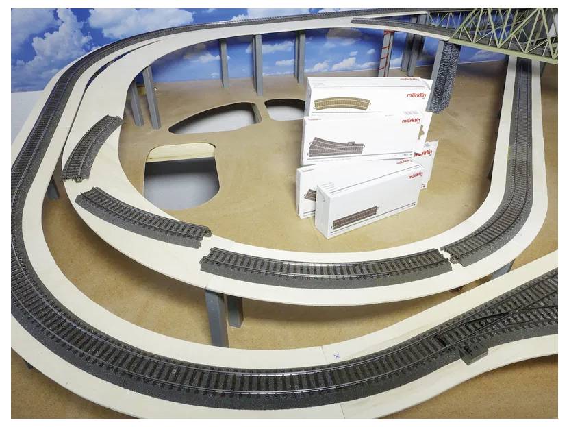 Model train tracks set up on a wooden base, with several stacks of unopened track boxes nearby, suggesting assembly or construction.