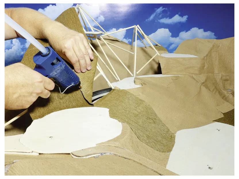 A person uses a glue gun to attach brown paper to a structure made of sticks, resembling a miniature landscape with a blue sky backdrop.
