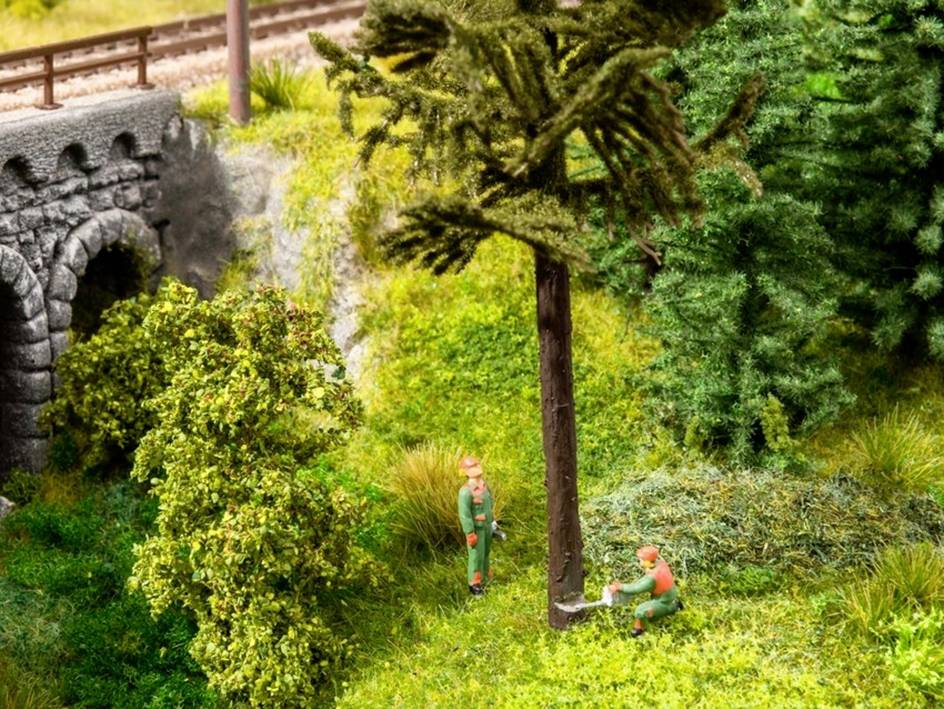 Two miniature figures are felling a tree in a modelled forest beside a railway bridge.