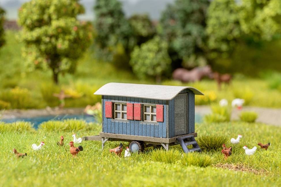 A blue, small farmhouse with red shutters sits on wheels in a green meadow. Surrounded by chickens and trees.