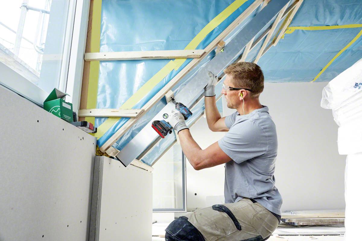 A construction worker is securing an insulation membrane to a roof pitch. He is wearing safety gloves and ear protection.