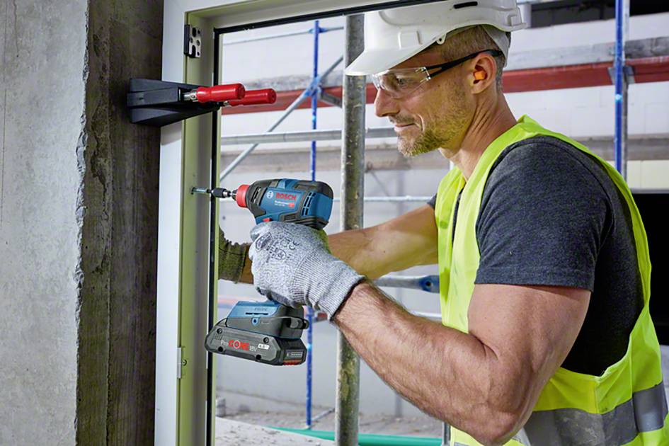 A construction worker in a high-visibility vest and hard hat is installing a window using a cordless drill. A scaffolding is visible in the background.
