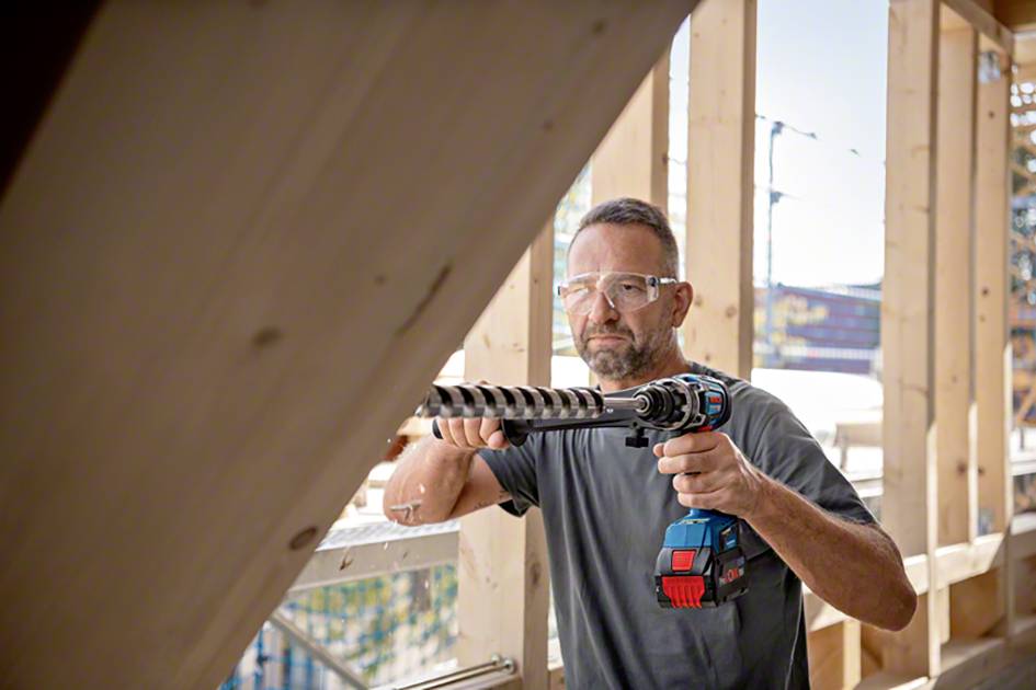 A man is wearing safety glasses and holding an electric drill in a wooden building, possibly during construction work.
