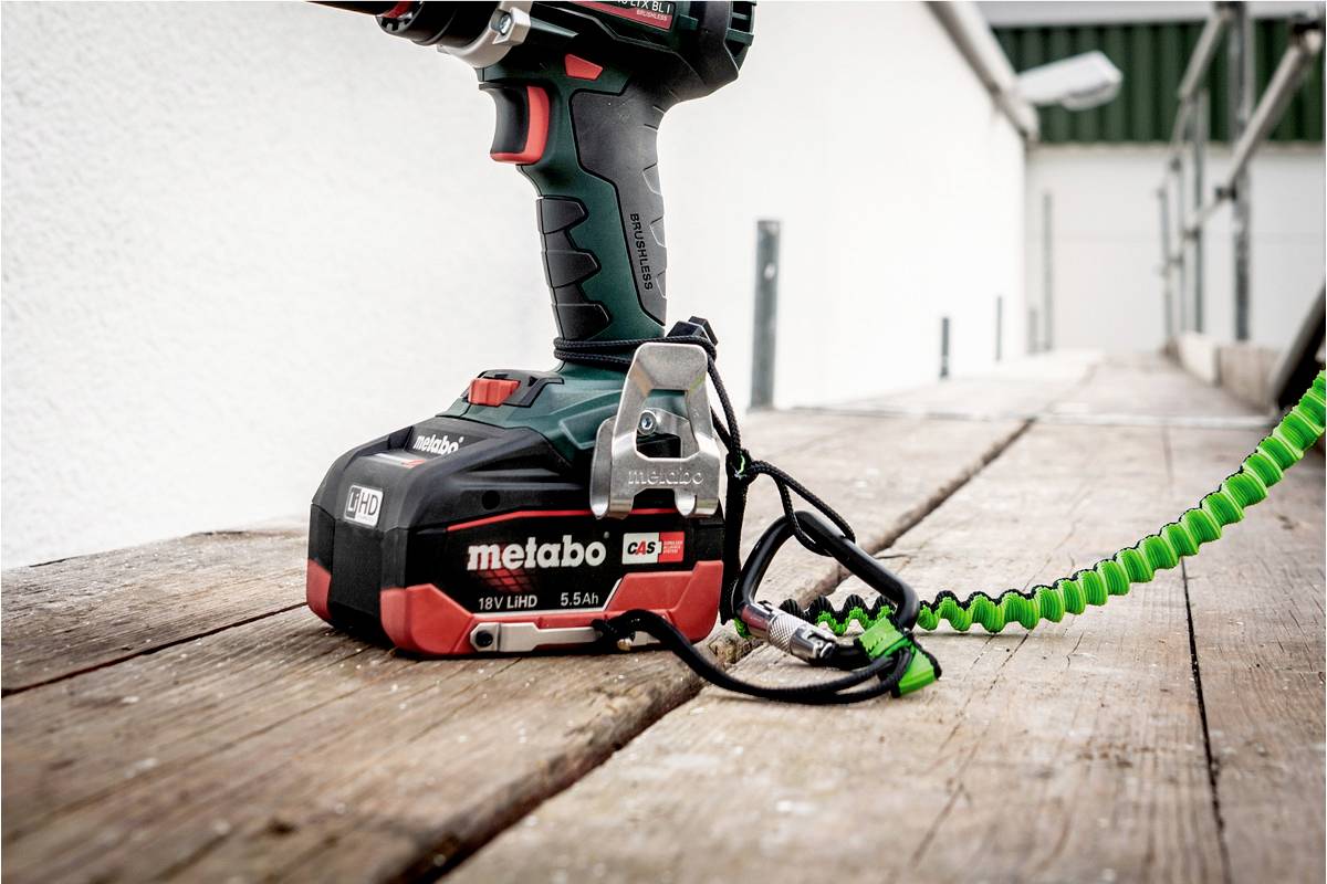A Metabo cordless drill with a green spiral cord lies on a wooden board outdoors.