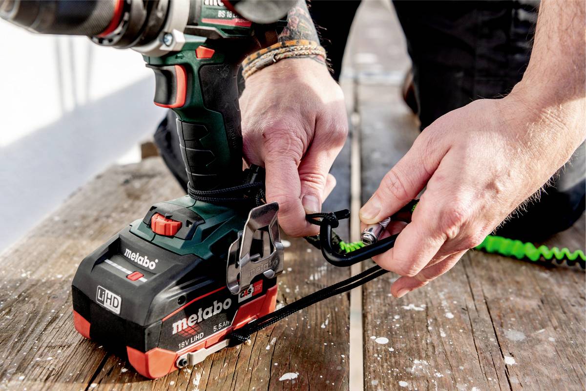 A person secures a safety line to a drill on a wooden floor. The drill is a Metabo model.