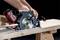 A hand operates a circular saw cutting through a wooden board. The saw is black with a silver blade and red battery.