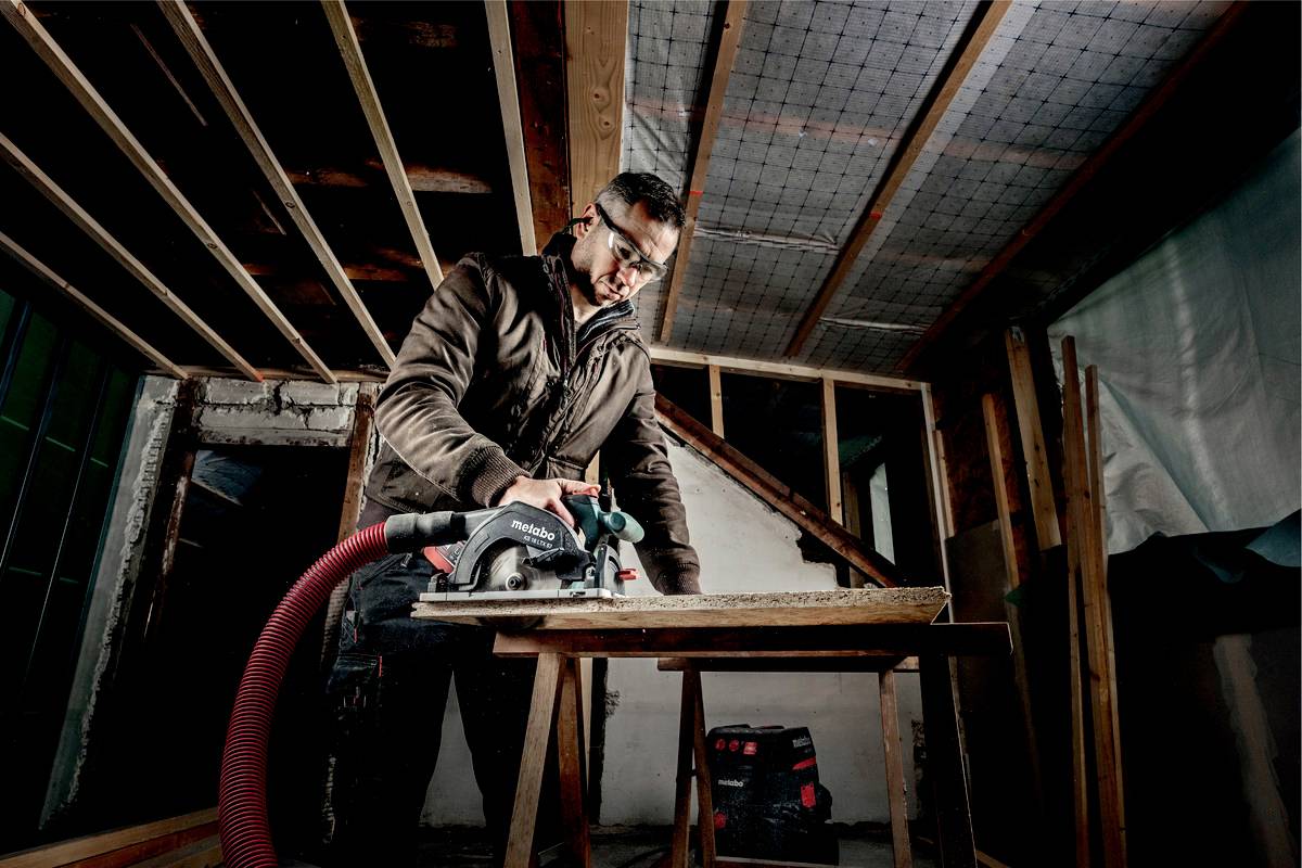 A man in protective clothing is using an electric saw to cut a wooden board on a building site. Wooden beams can be seen in the background.