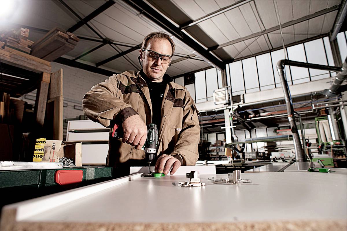 A worker in a workshop is wearing safety glasses and using a drill to assemble parts on a white board.