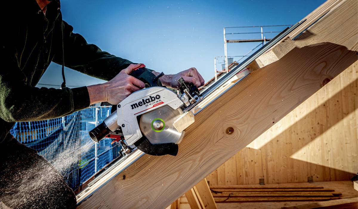 A person is working with a circular saw on a wooden scaffold in daylight. Wood shavings are flying from the saw.