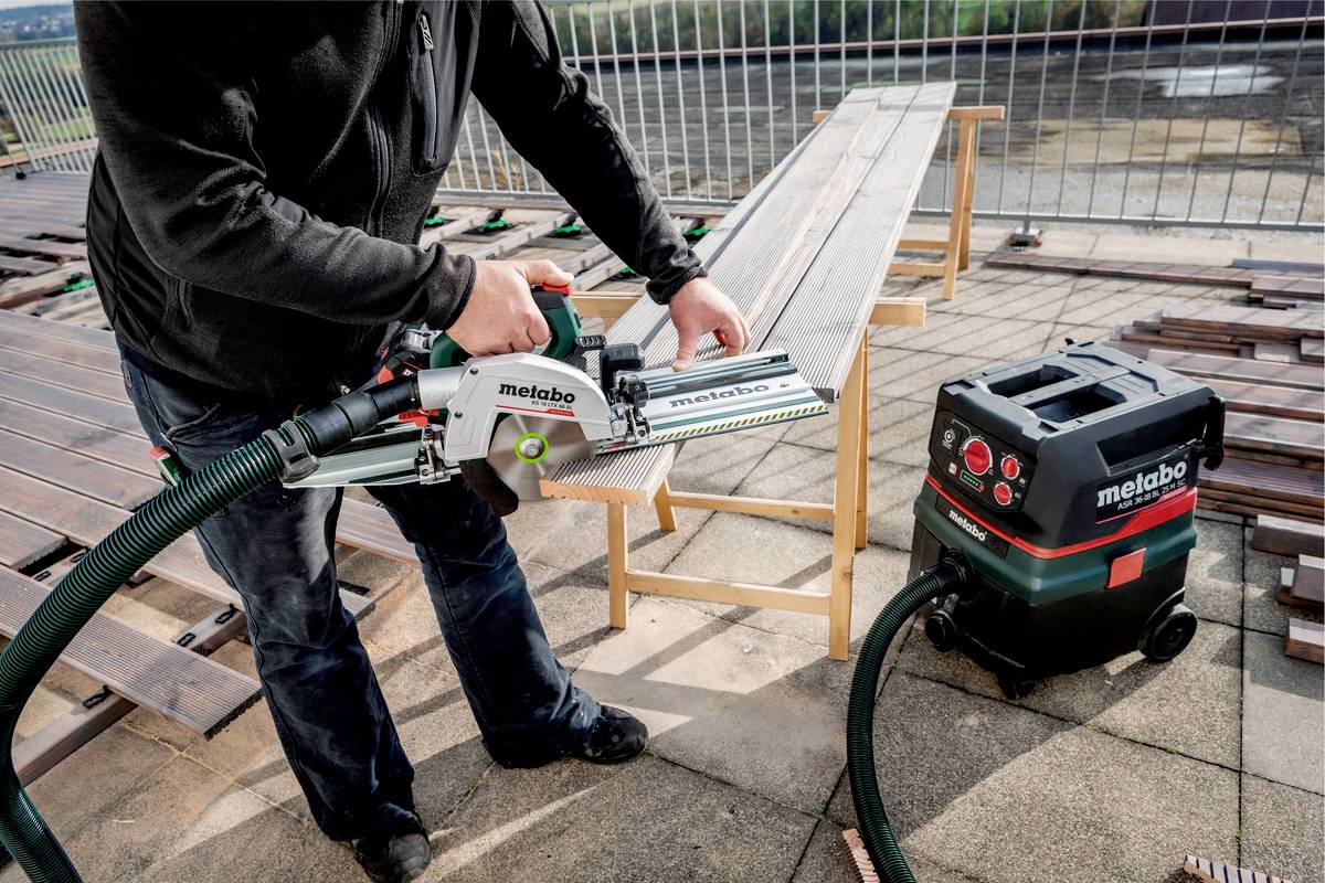 A person is working with a Metabo mitre saw on a terrace. A vacuum cleaner is connected for dust extraction. The environment appears industrial.
