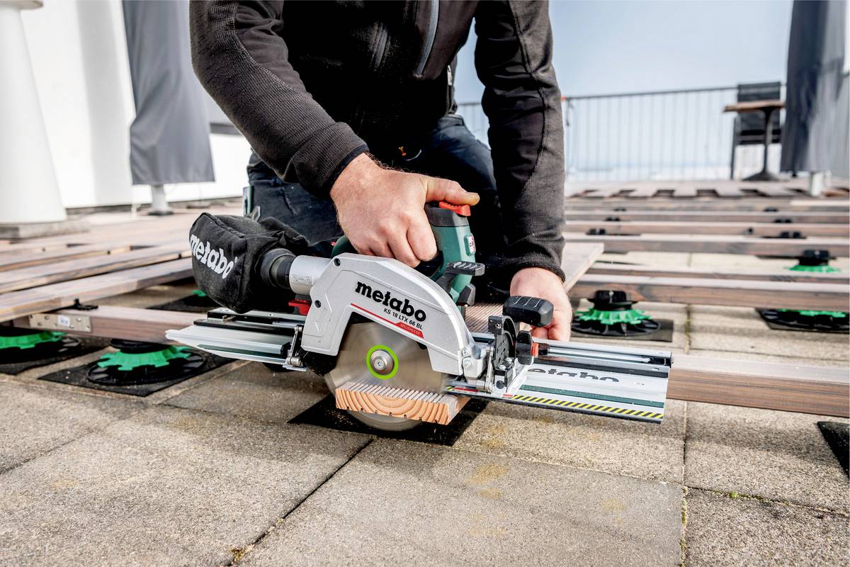 A person is sawing wooden boards on a terrace using a Metabo circular hand saw. Further boards and a balustrade are visible in the background.