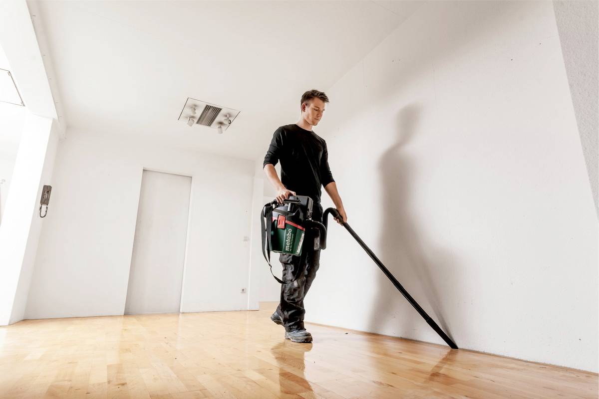 A person is hoovering an empty room with a wooden floor. The walls are white, and the room is brightly lit.