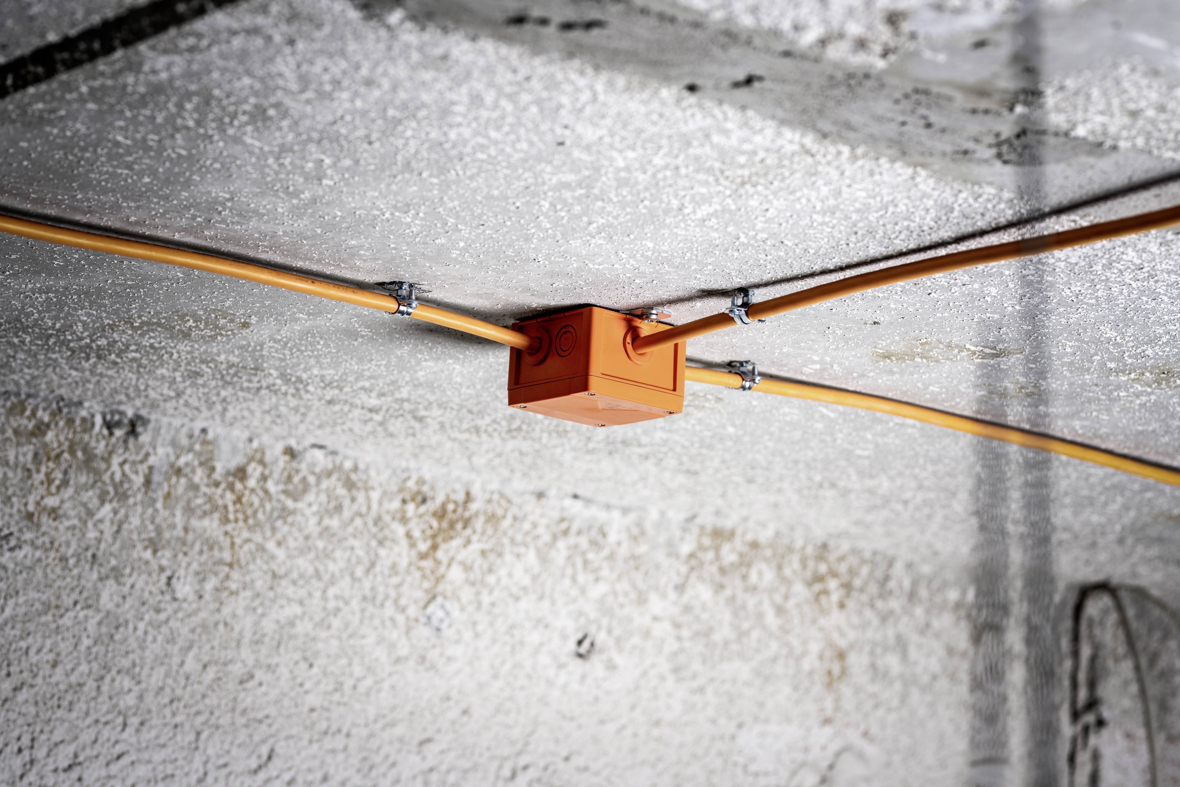 Orange distribution box mounted on a ceiling, connected by cables in four directions. Concrete background.