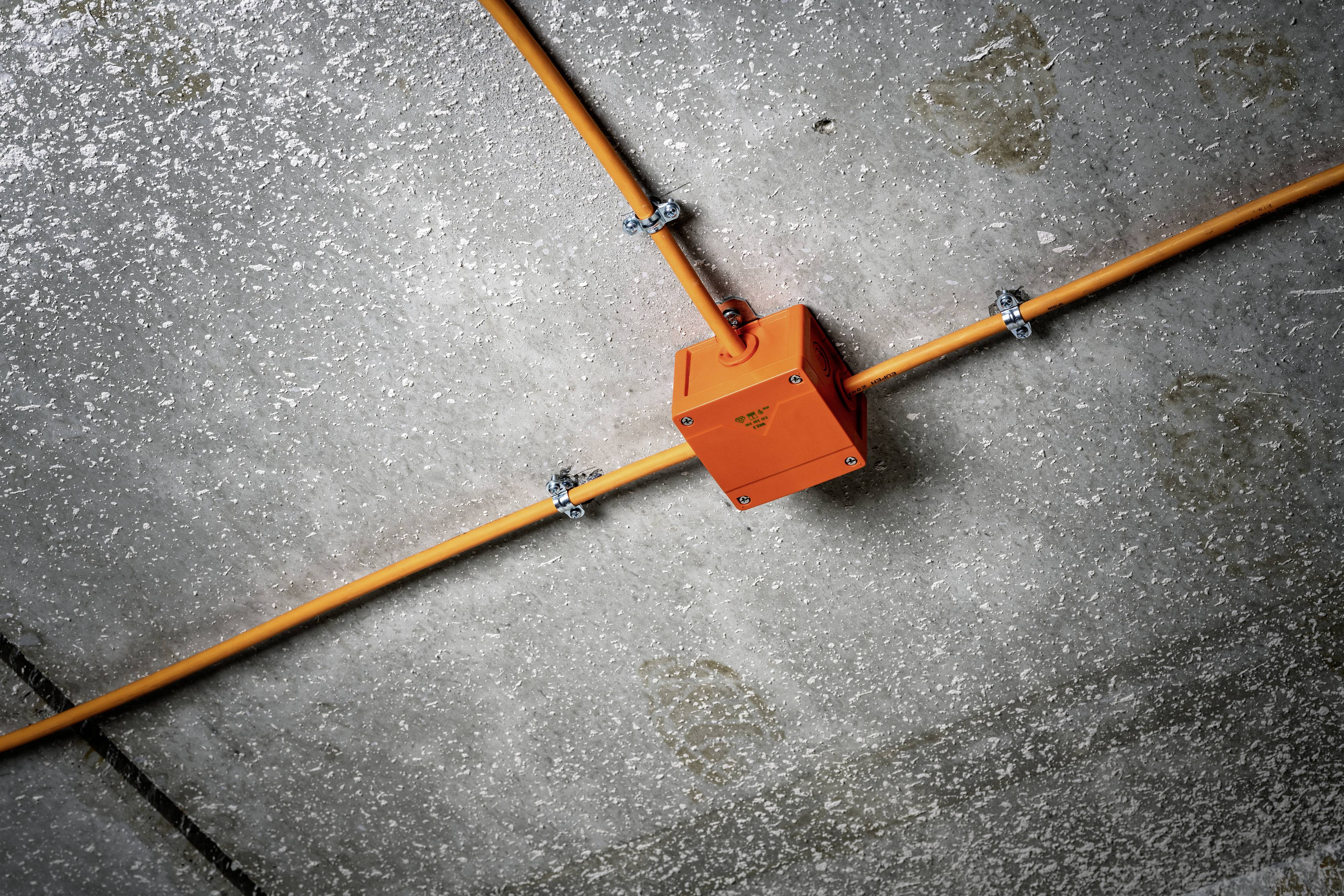 An orange junction box with cables is mounted on a grey concrete wall. It is positioned facing downwards to the left.