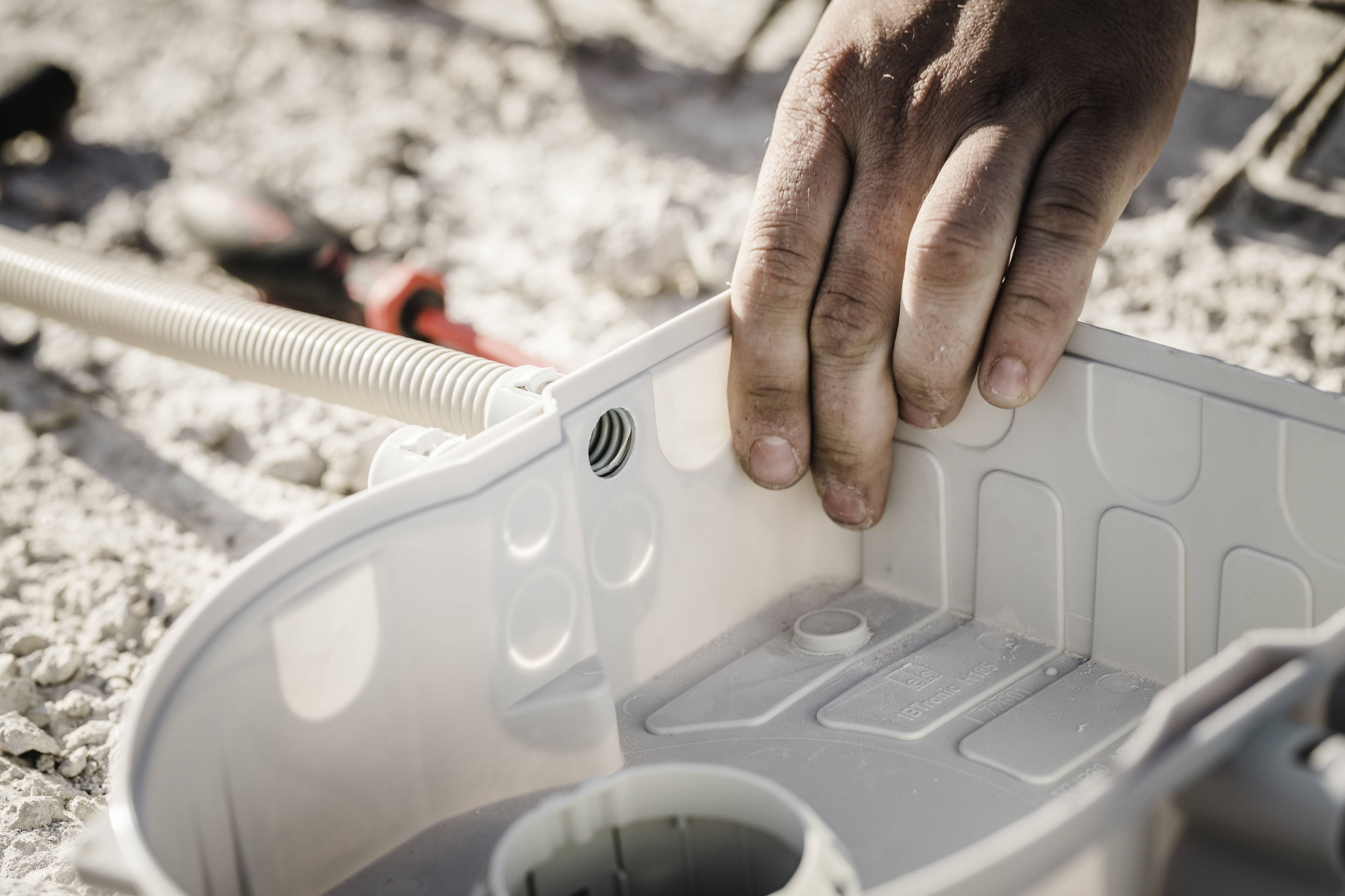 Hand mounting a grey plastic housing component on an unspecified surface. Blurred tools visible in the background.