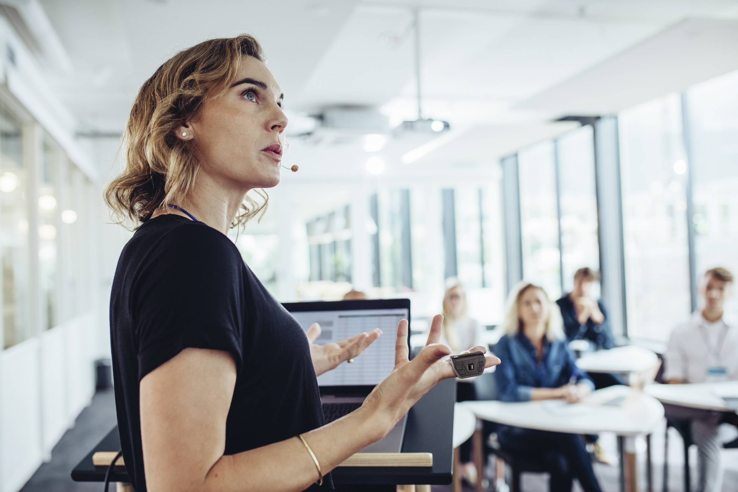 A woman with a microphone is giving a presentation in a modern conference room to an attentive audience.