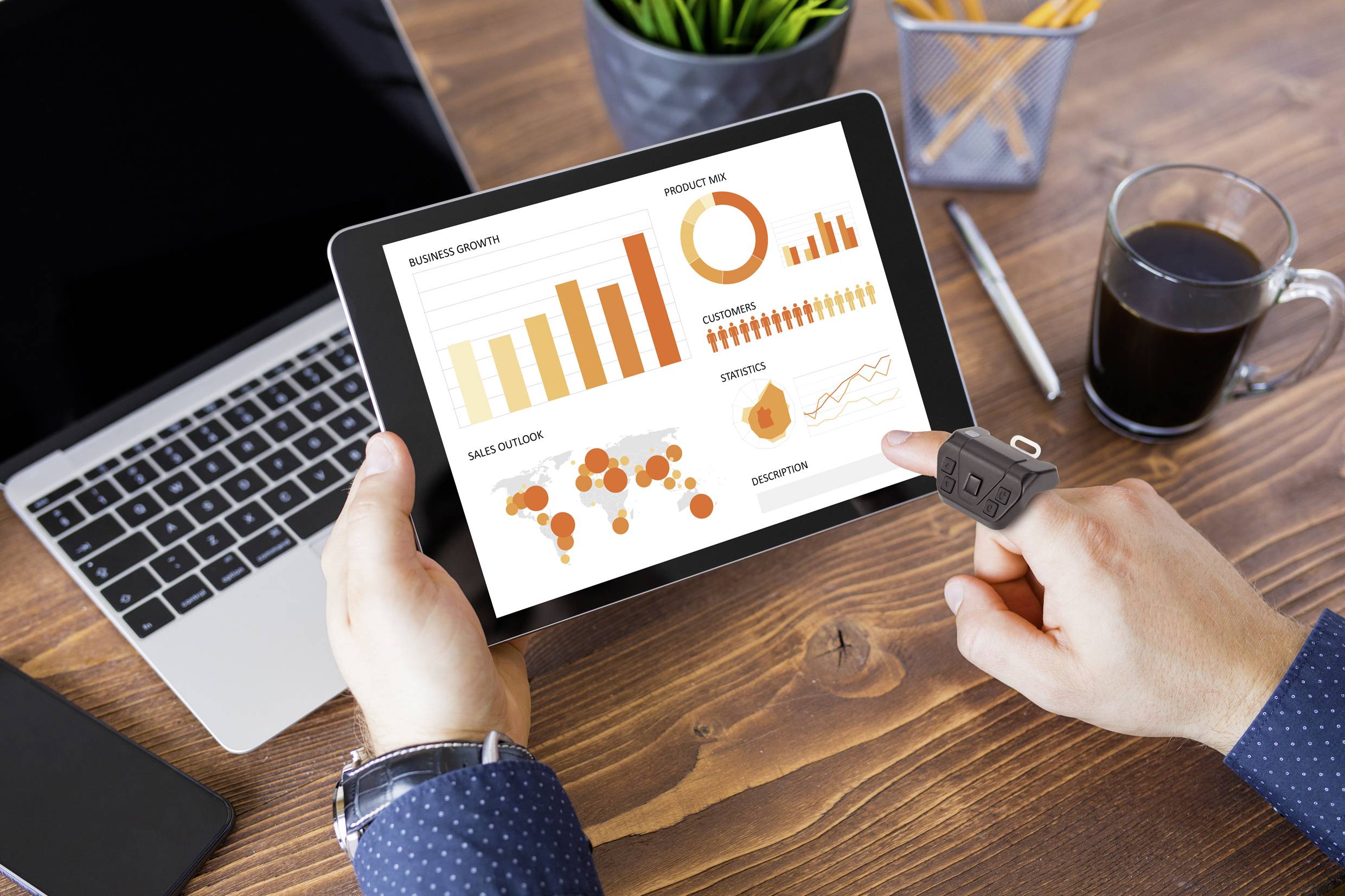 Hand holding a tablet displaying a dashboard with graphs: a bar chart showing revenue growth, a pie chart representing market shares, and a world map with sales figures. Laptop and coffee in the background.