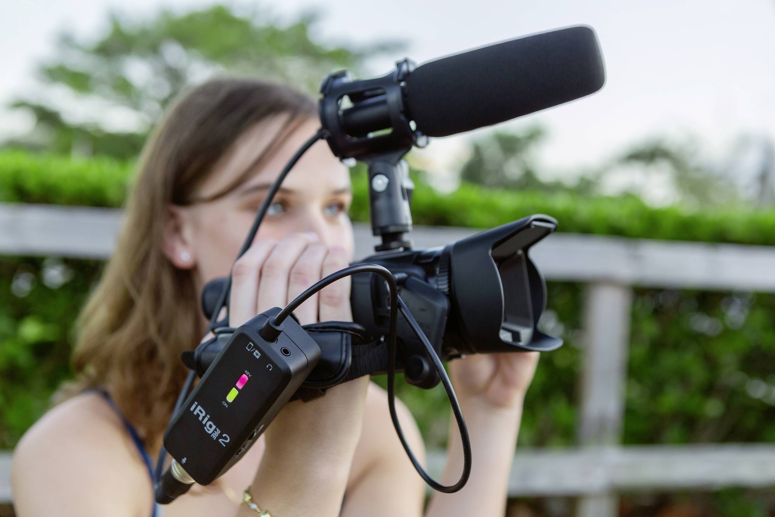 A person is filming outdoors with a video camera, equipped with a microphone and an iRig2 audio interface. Trees are visible in the background.
