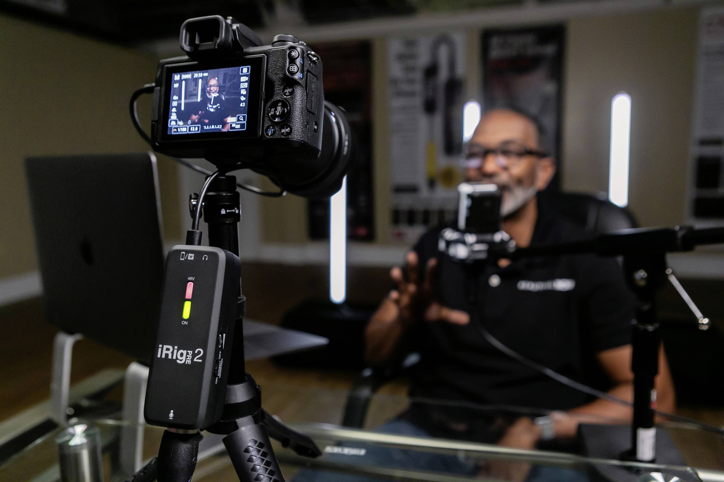 A man in a studio is speaking into a microphone while being filmed by a camera. A laptop is positioned to his left.