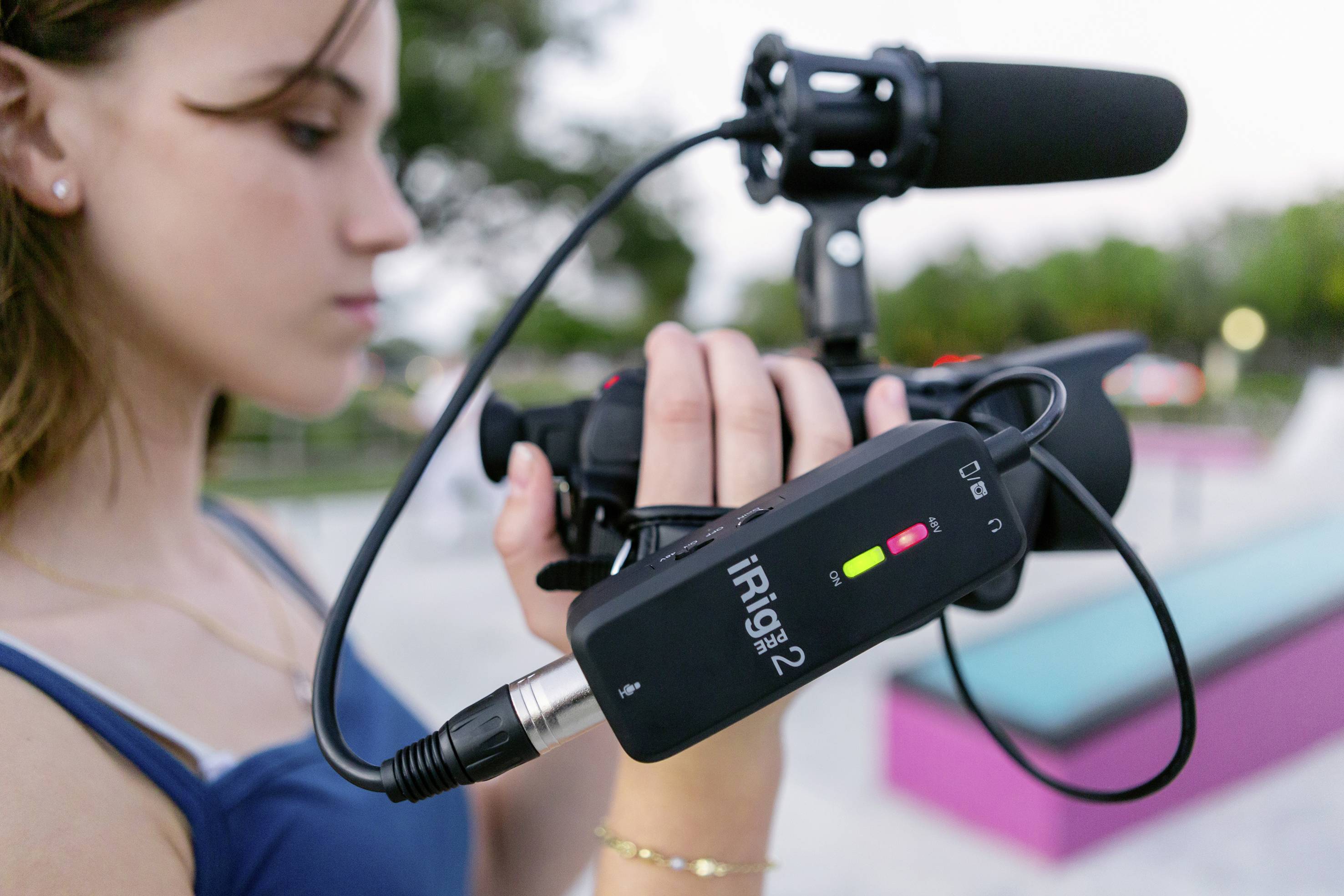 A person is holding a camera with a microphone and an iRig device for sound recording. A blurry park is visible in the background.