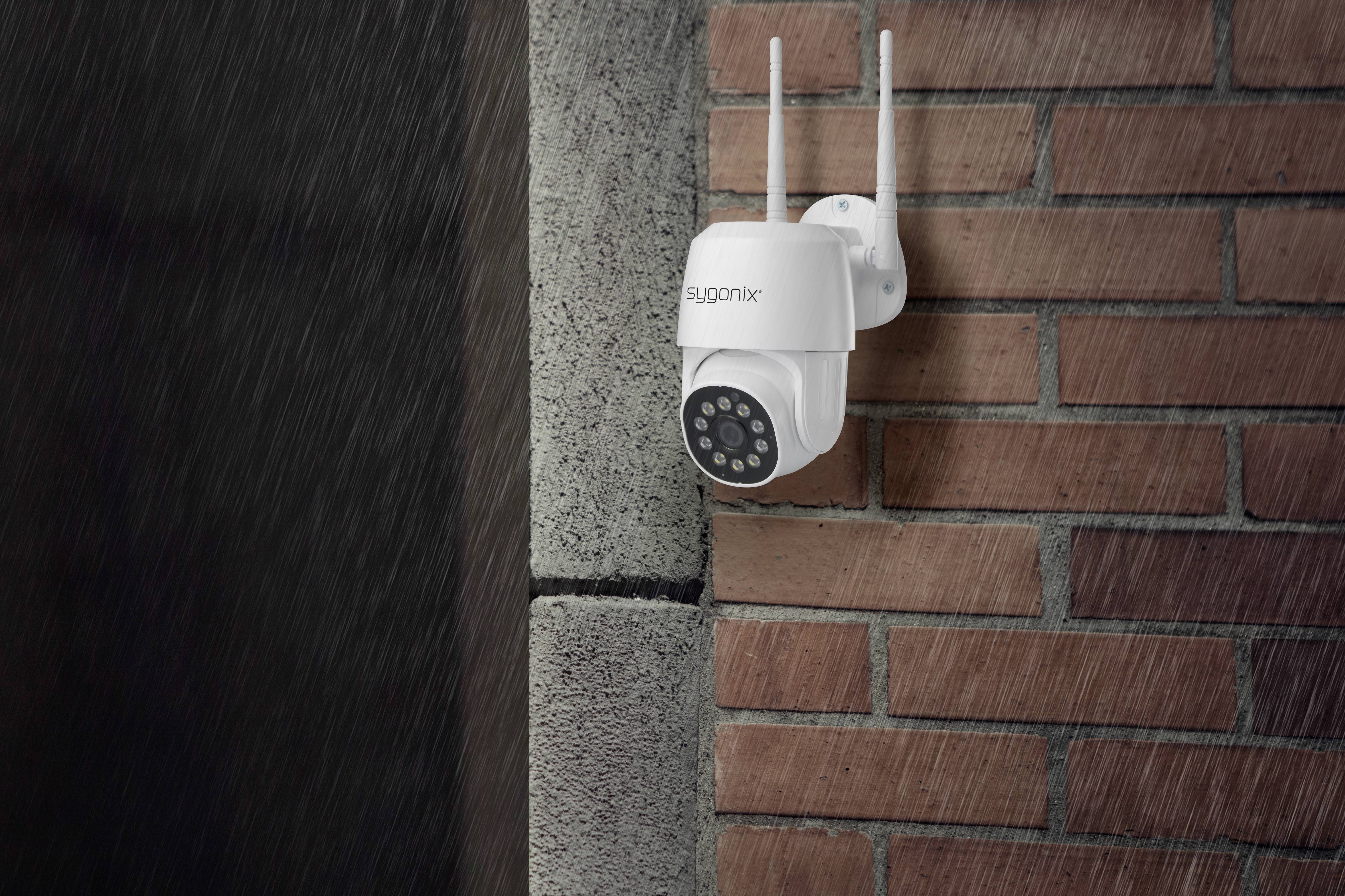 A surveillance camera is mounted on a brick wall outside in the rain. It has two aerials and a dark lens.