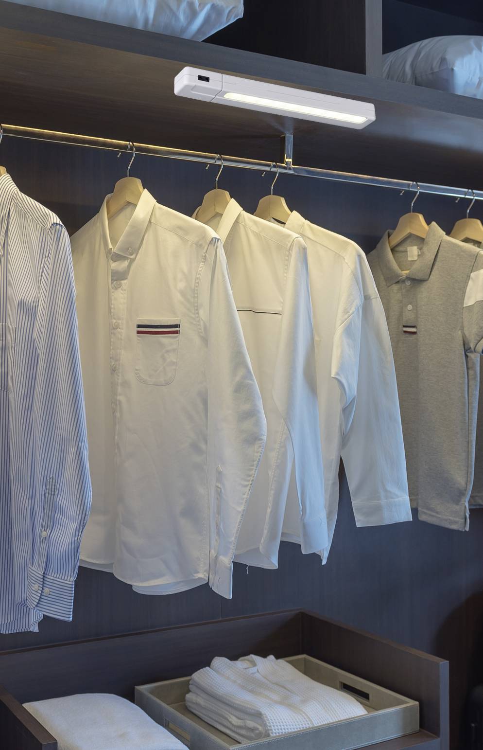 White and striped shirts hang neatly on wooden coat hangers in an illuminated wardrobe. Folded towels are placed underneath.