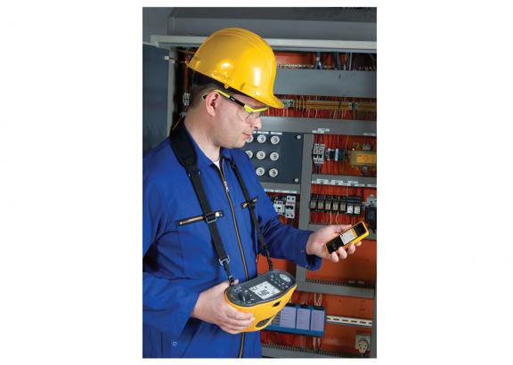 An electrician in blue workwear and a yellow safety helmet is checking measuring instruments in front of an electrical control cabinet with electrical components.