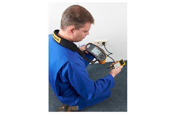 A technician in blue workwear is testing electrical equipment with a measuring device and recording the results on a handheld device.