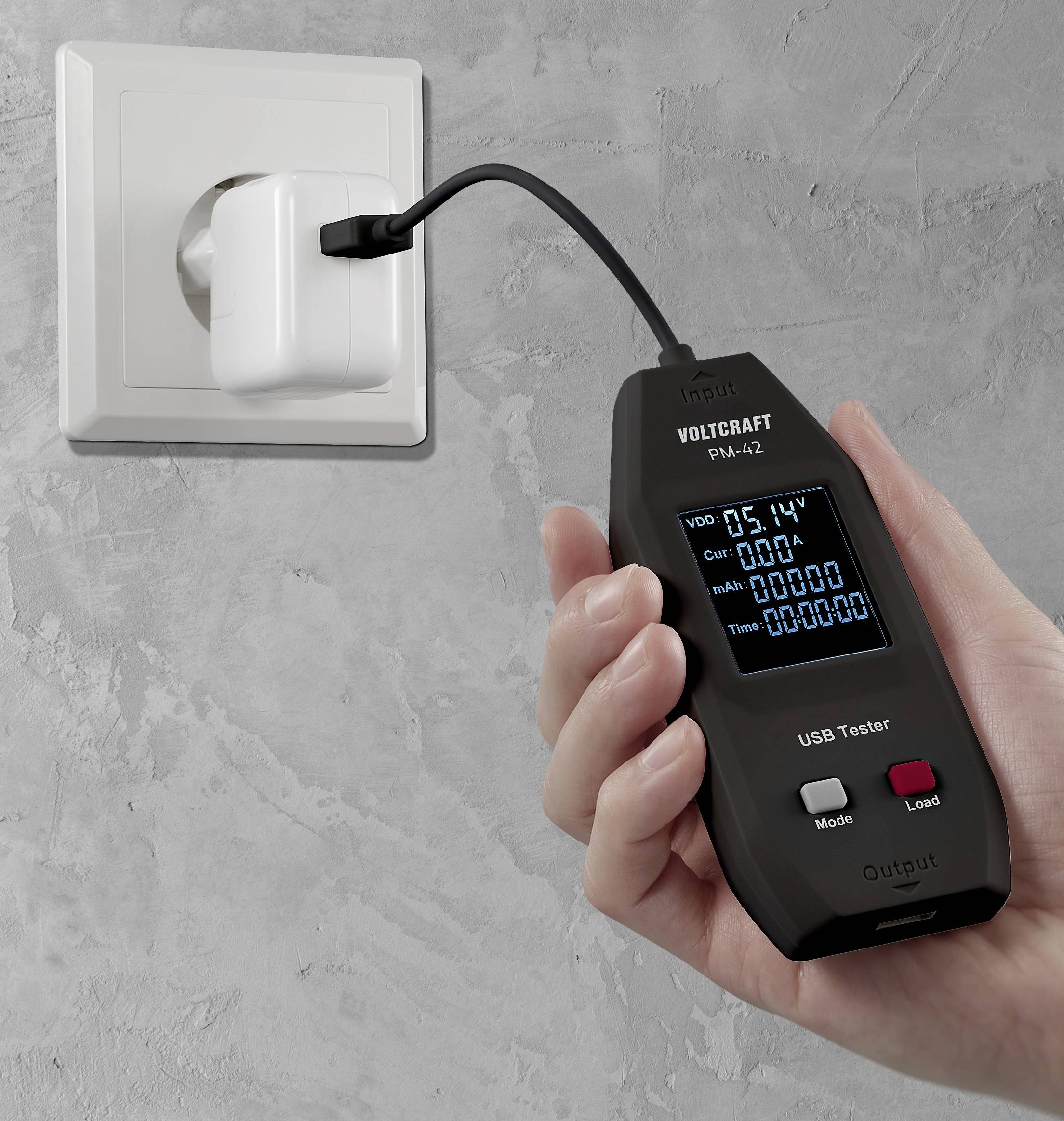 A hand is holding a USB tester plugged into a charger at a wall socket. The screen displays various measurement readings.