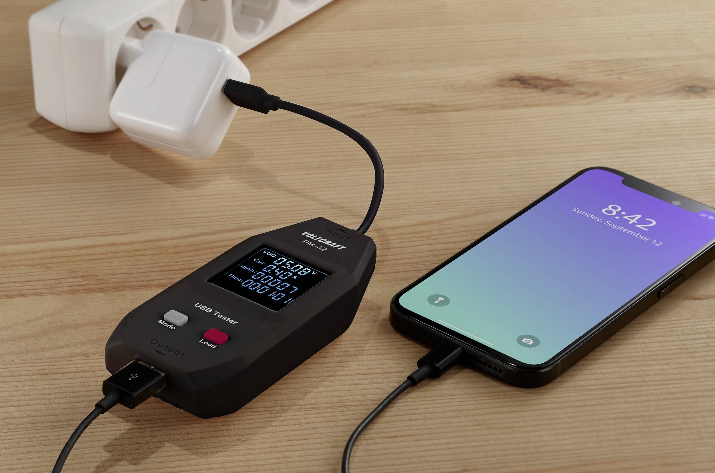 A smartphone and a USB power metre are connected on a wooden table. The display shows voltage, current, and power.