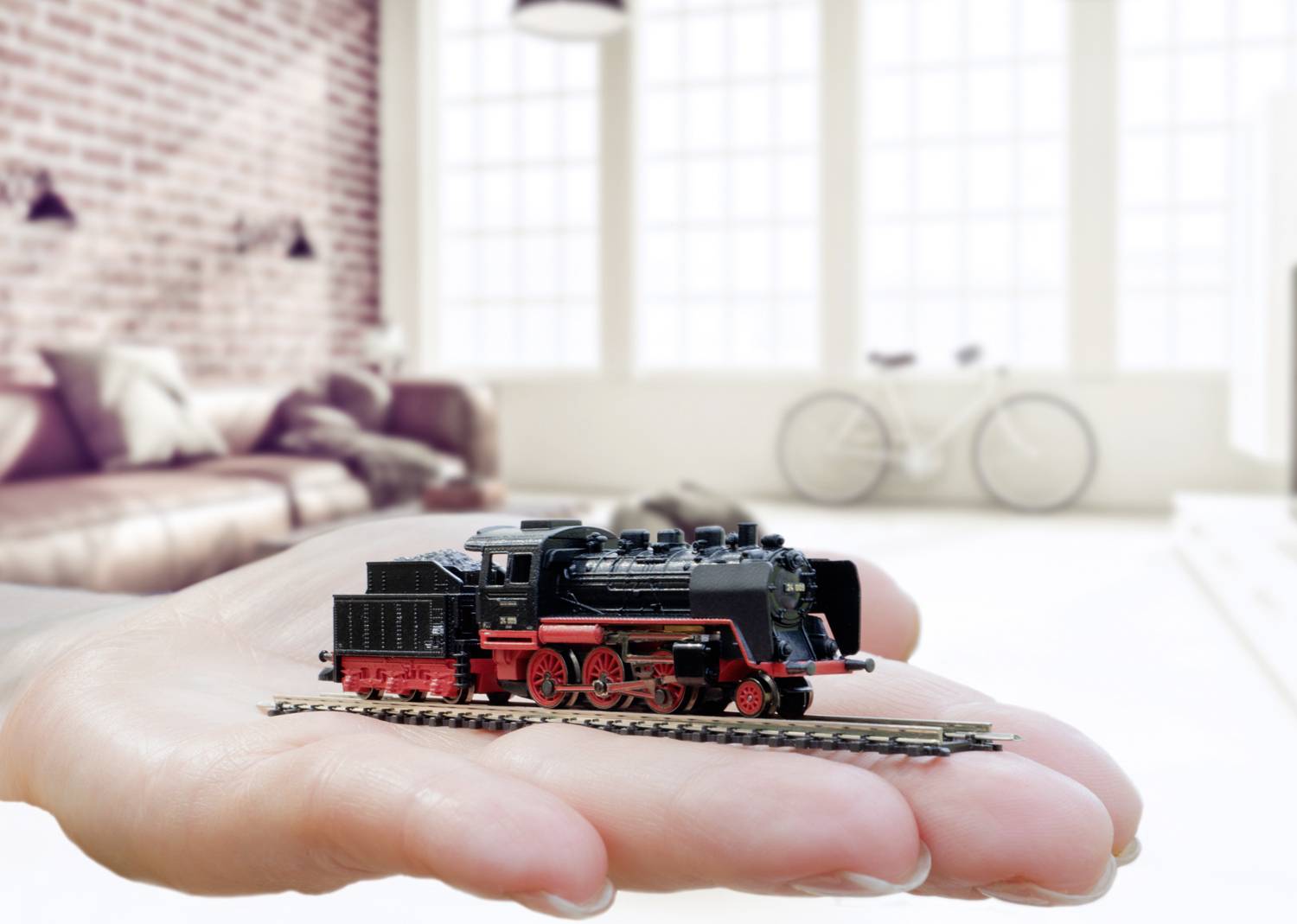 A hand is holding a model steam locomotive with a red chassis and black boiler. A modern living room is visible in the background.