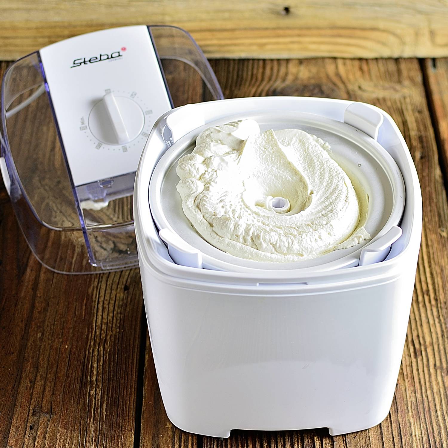 A white ice cream maker, currently preparing ice cream, is standing on a wooden table. The lid is open, and the ice cream is almost ready.