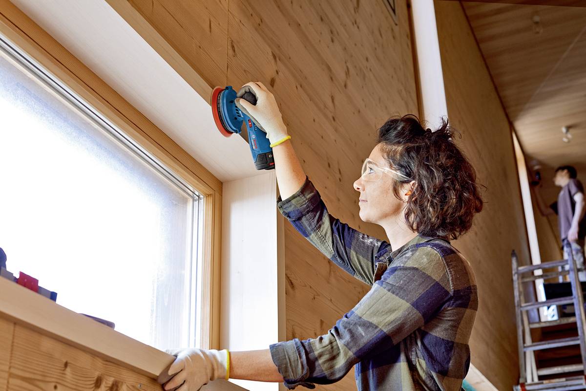 A person is sanding the wooden window frame with an electric sanding tool. They are wearing safety glasses and gloves.