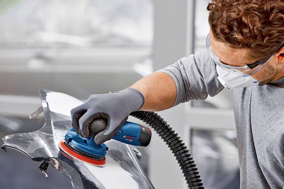 A person is grinding a metal surface with an electric grinding tool. They are wearing safety glasses, a mask, and gloves.