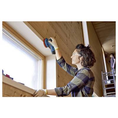 A person using a power sander on a wooden window frame in a well-lit room. They are wearing safety goggles and gloves.