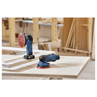 Two cordless power sanders on a workbench in a woodshop, with wooden shelves and tools in the background.
