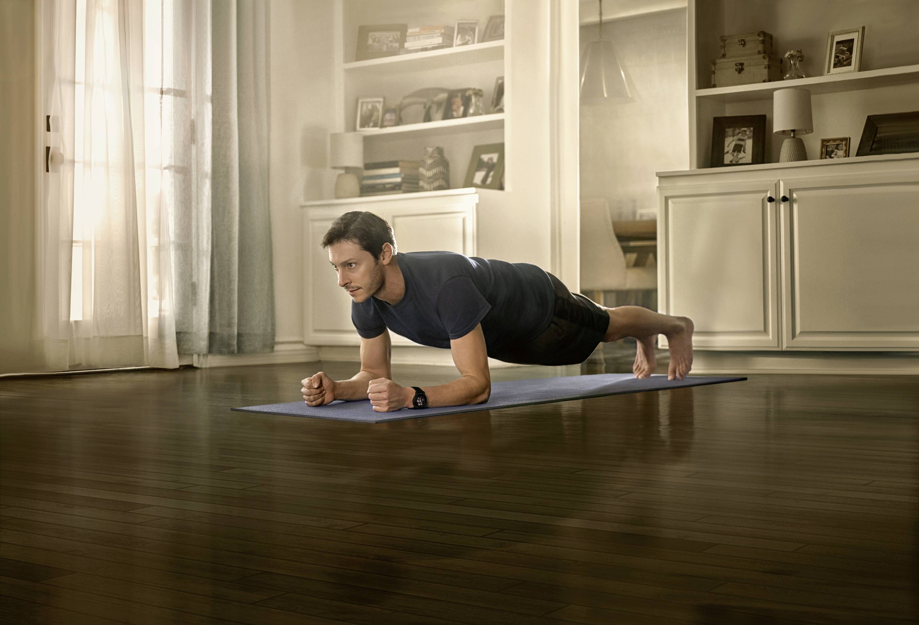 A man is performing a forearm plank on a yoga mat in a living room. Cupboards and windows are visible in the background.