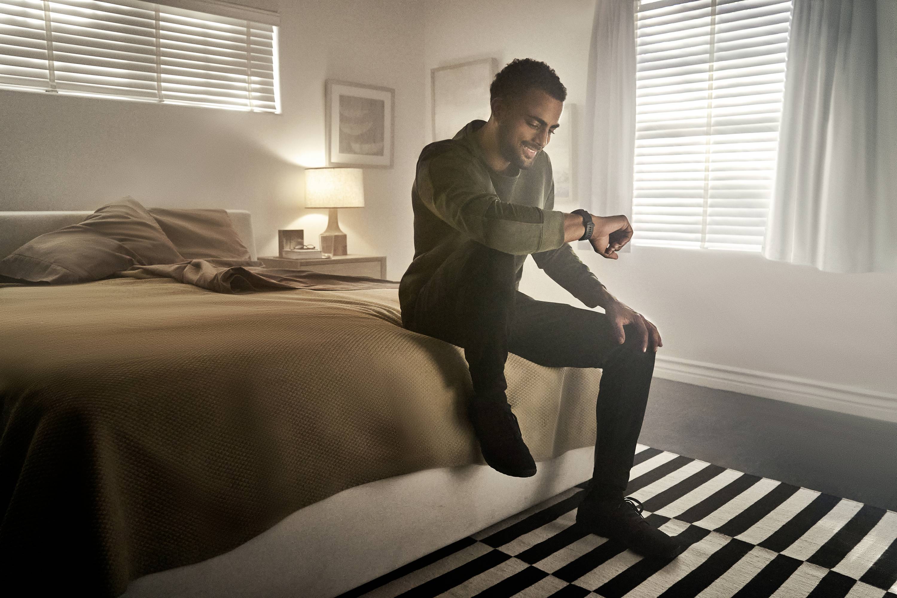 A man is sitting smiling on a bed, looking at his smartwatch. In the background, there is a bedside table with a lamp and a window.