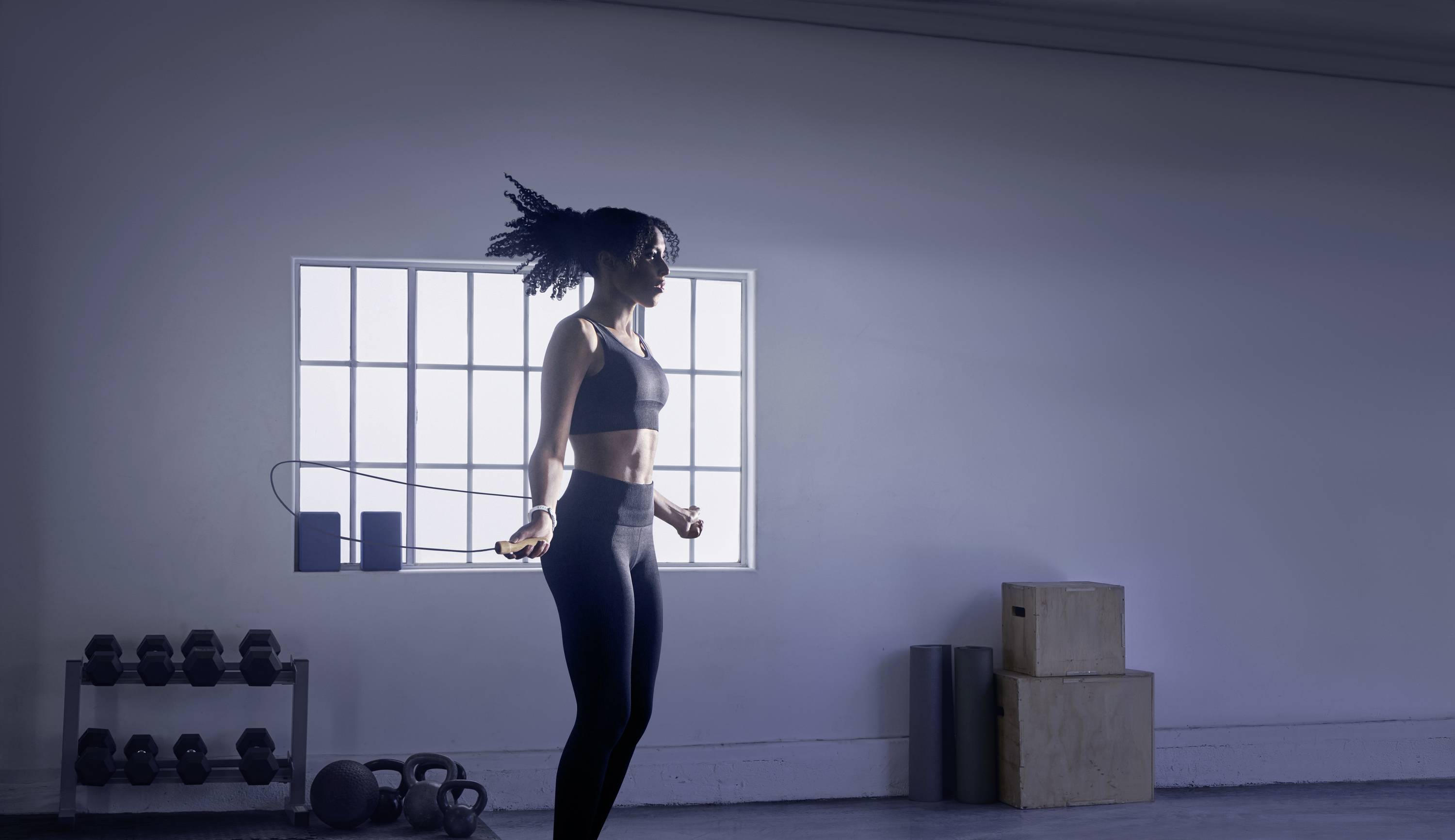'Woman skipping rope in a minimalist fitness studio with dumbbells and boxing equipment in the background under natural light.'
