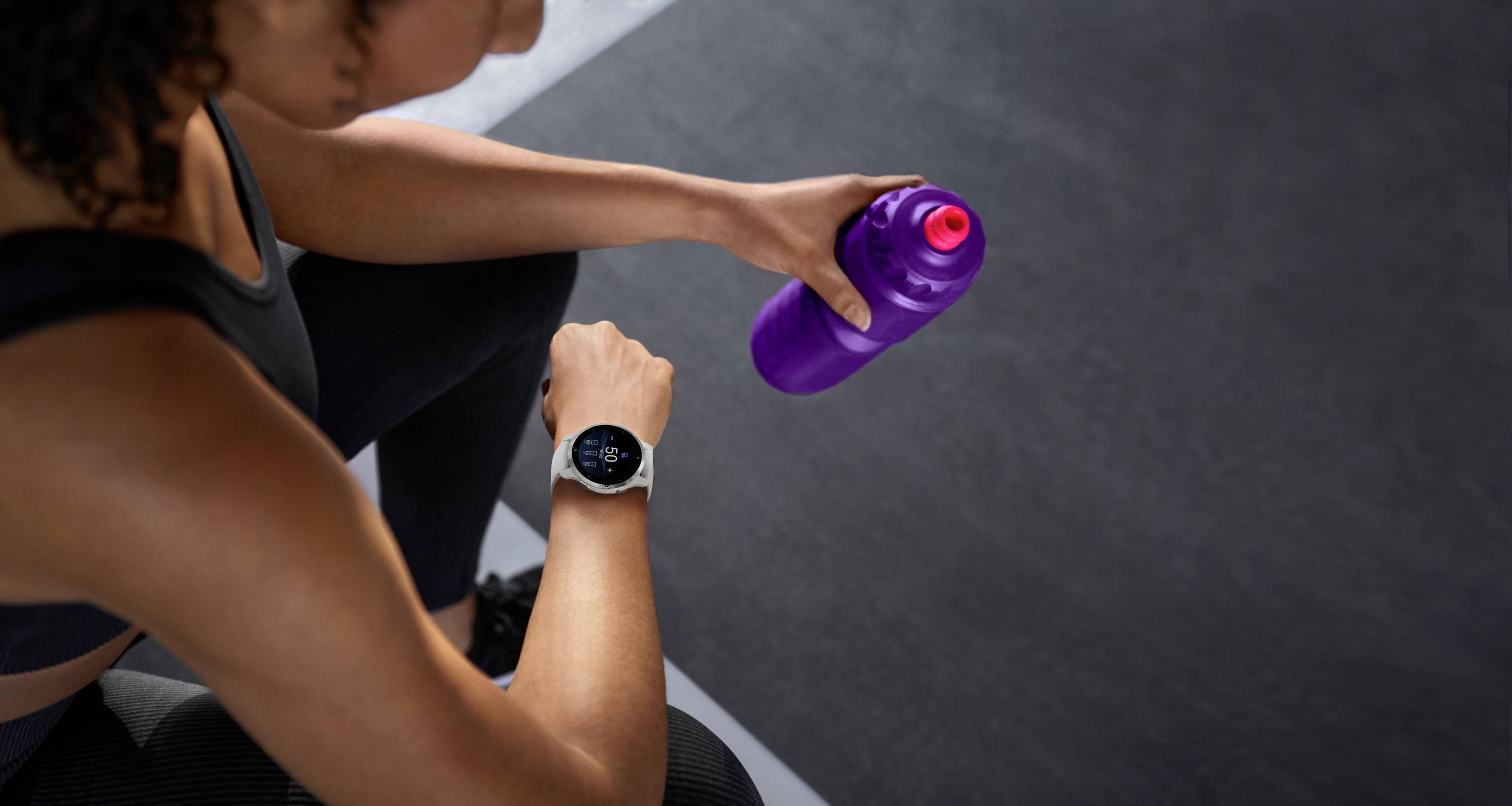 A woman is holding a purple water bottle in her hand. She is wearing sports clothing and a black smartwatch.