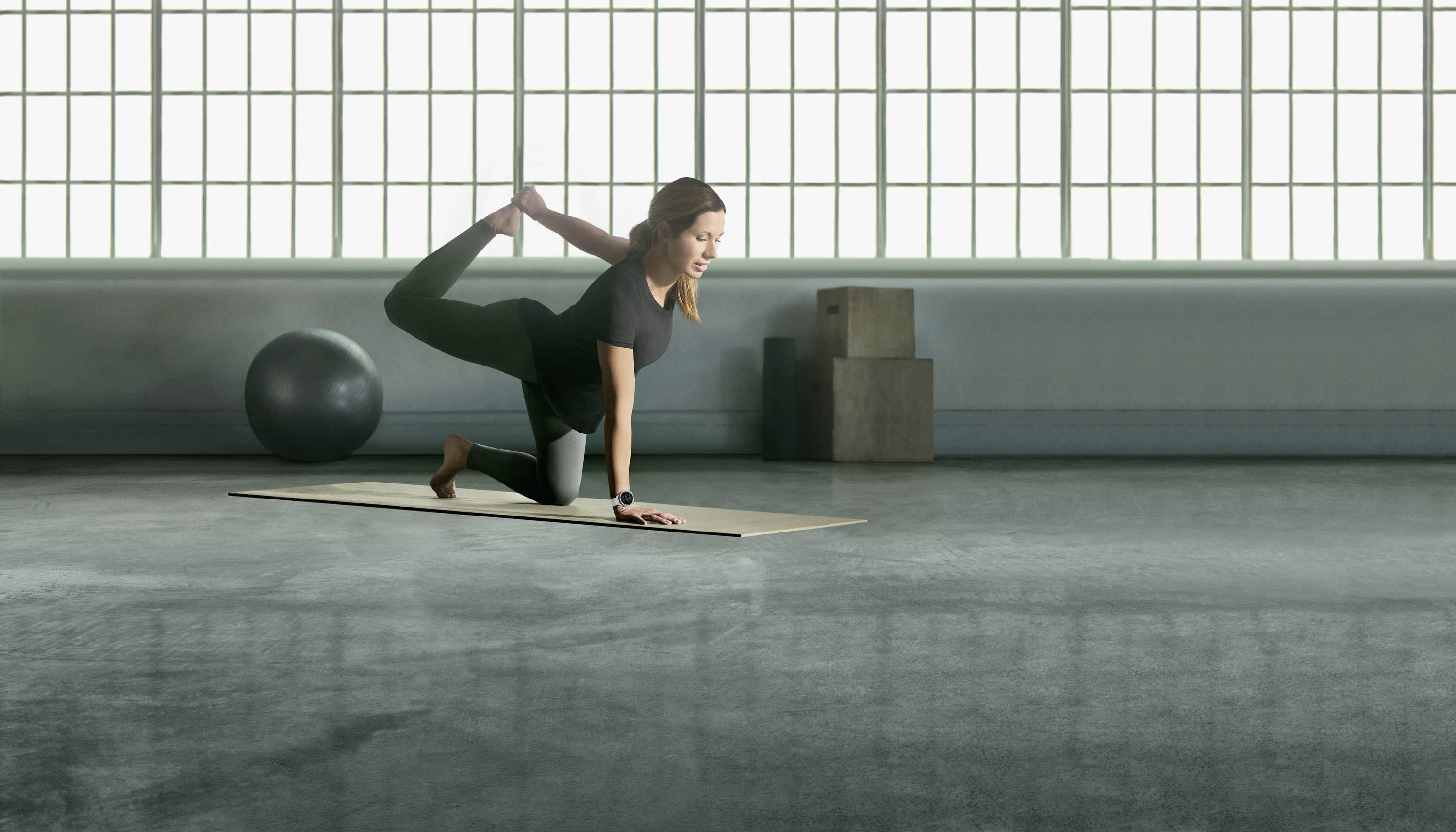 A woman is practising yoga in a studio. She is balancing on a mat in a four-legged pose, with one leg bent upwards. In the background: an exercise ball.