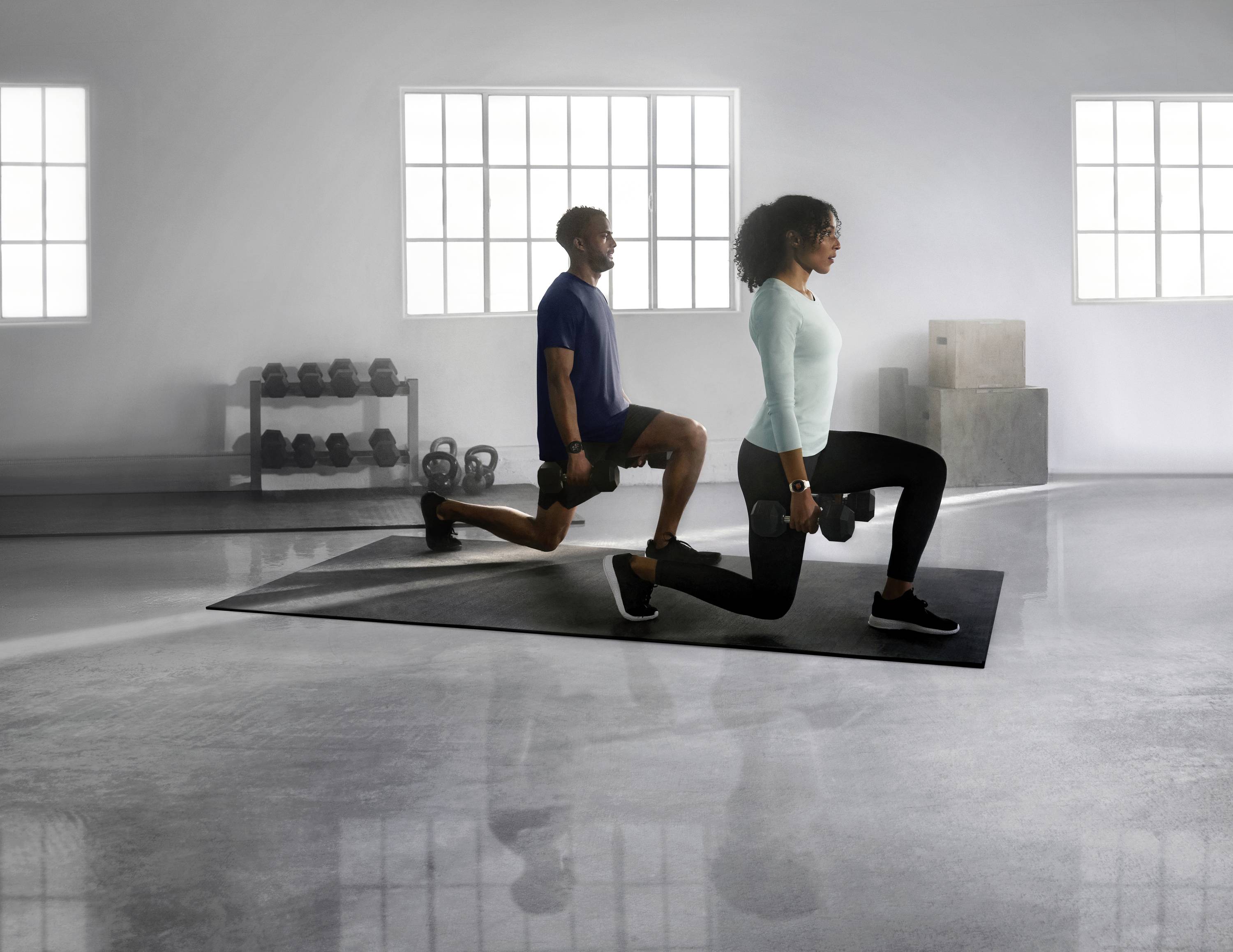 A man and a woman are training with dumbbells in a gym. Both are performing lunges on a black mat. Dumbbells and weights are visible in the background.