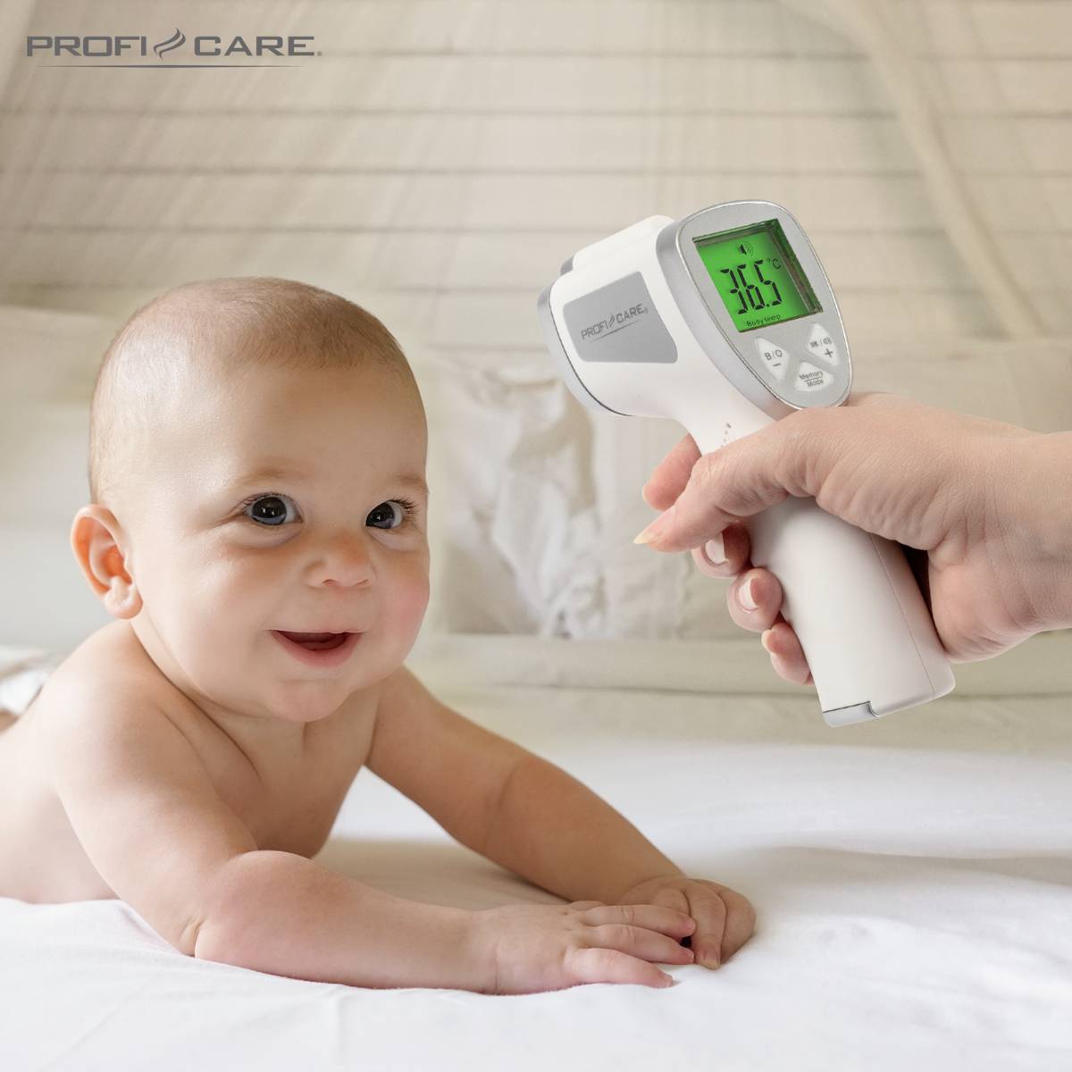 A baby is lying on its tummy and smiling. An infrared thermometer is held against the baby's forehead. The display shows 36.5°C.
