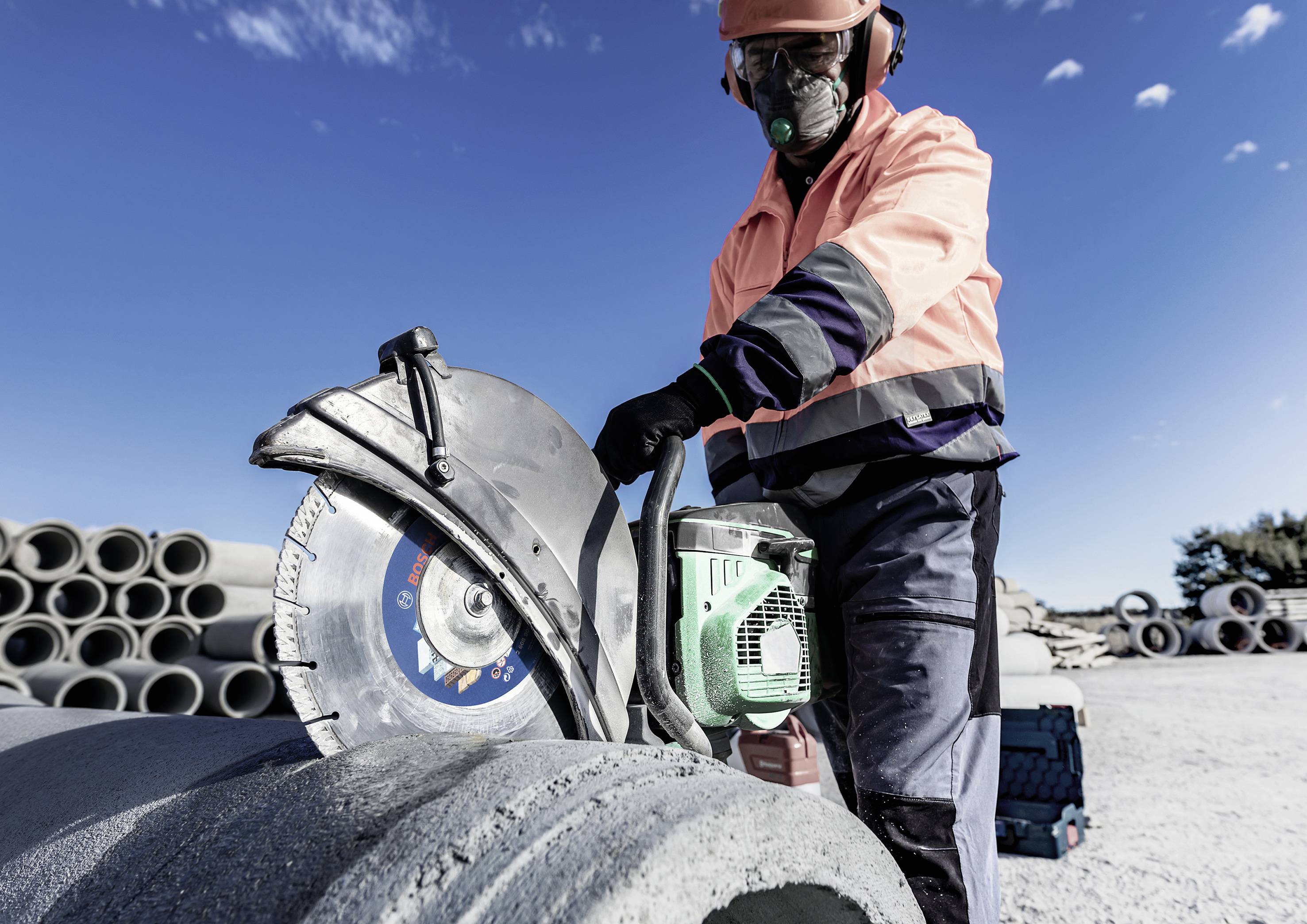 A construction worker is cutting concrete pipes with a chainsaw. Stacked pipes and a clear blue sky are visible in the background.